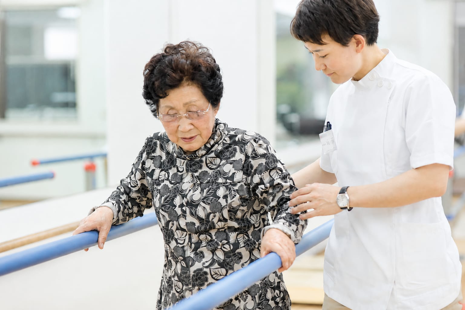 平行棒につかまり歩行訓練を行うご利用者様と付き添うスタッフ